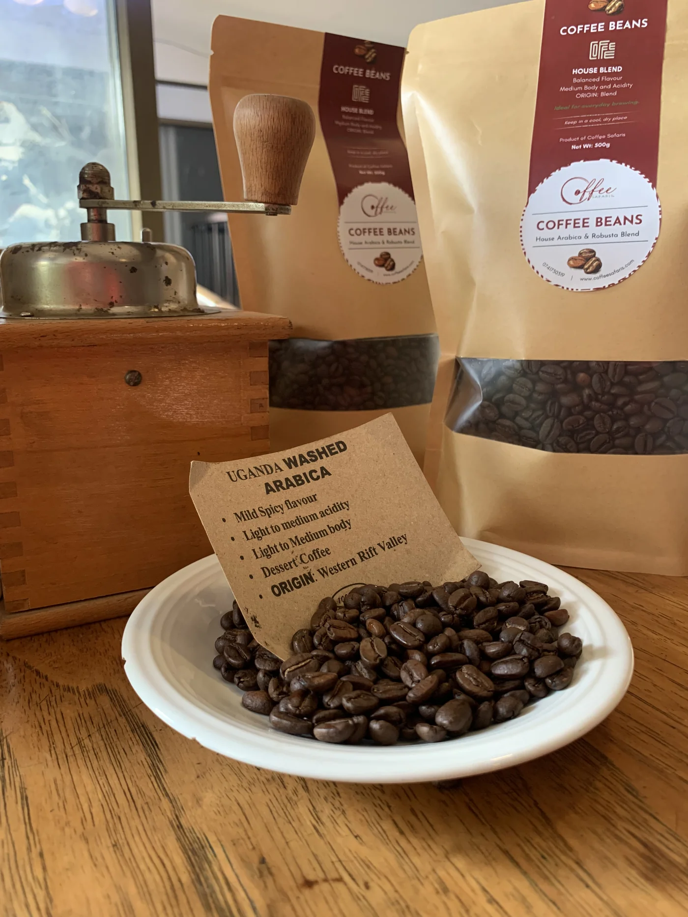 Coffee Safaris branded roasted bean bags, a plate of Uganda Washed Arabica beans with origin card, and a vintage hand grinder on a rustic wood table